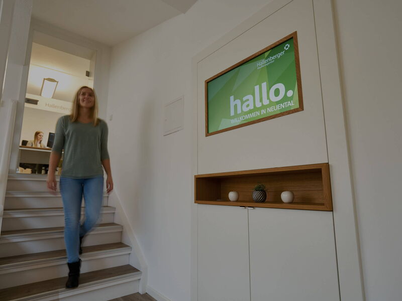 Eine Frau läuft eine Treppe hinunter, im Hintergrund ein Büroraum mit Frau am Schreibtisch, auf der rechten Seite hängt ein Bildschirm an der Wand mit einem Bild mit "hallo. Willkommen in Neuental".