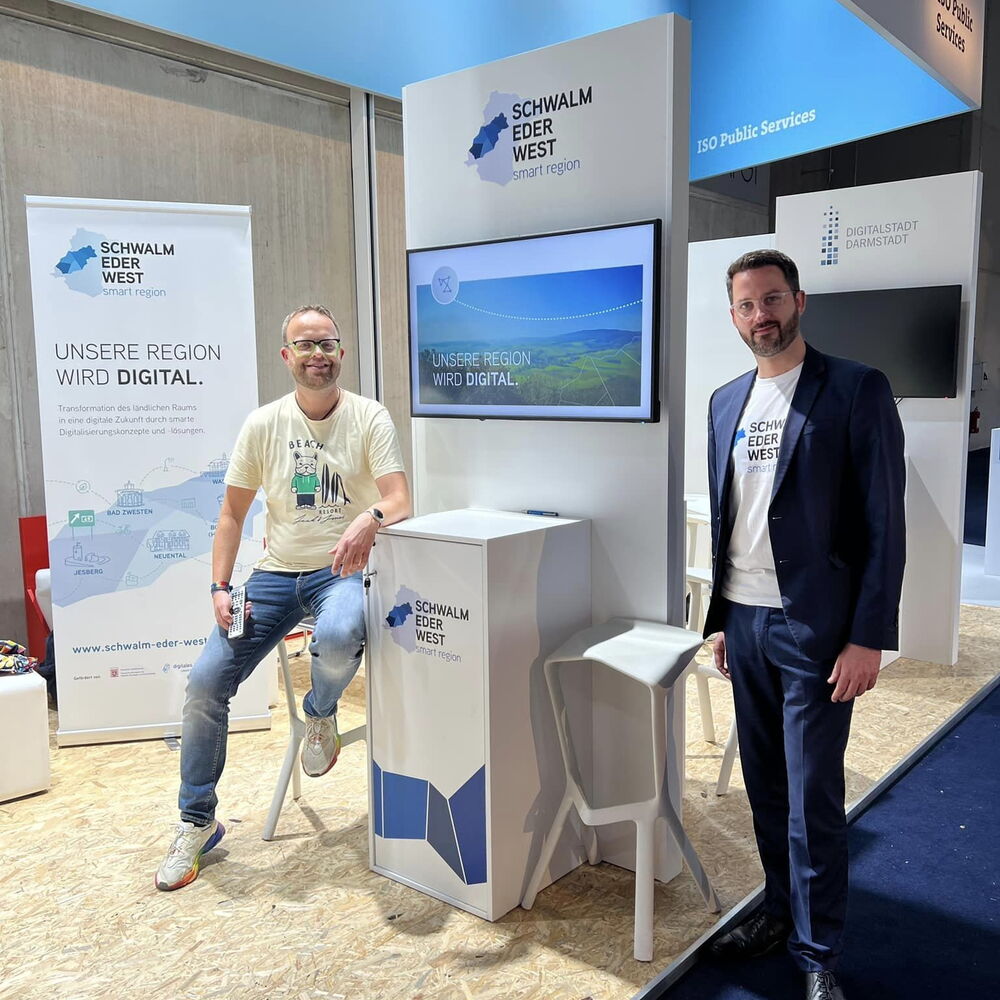 ein Messestand der Schwalm-Eder-West mit einem Fernseher und Bannern. Ein Mann sitzt auf einem Hocker und ein anderer Mann steht neben dem Messestand