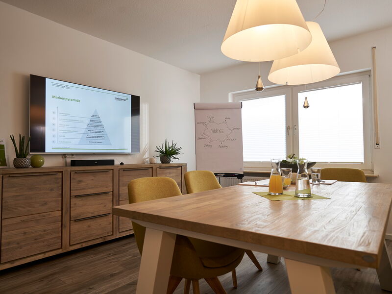 Ein Besprechungsraum mit Holztisch mit Getränken drauf, grüne Stühle drumherum, zwei Schirmlampen hängen von der Decke herunter, an der Wand hängt einer großer Bildschirm mit Bild, darunter steht ein Sideboard.