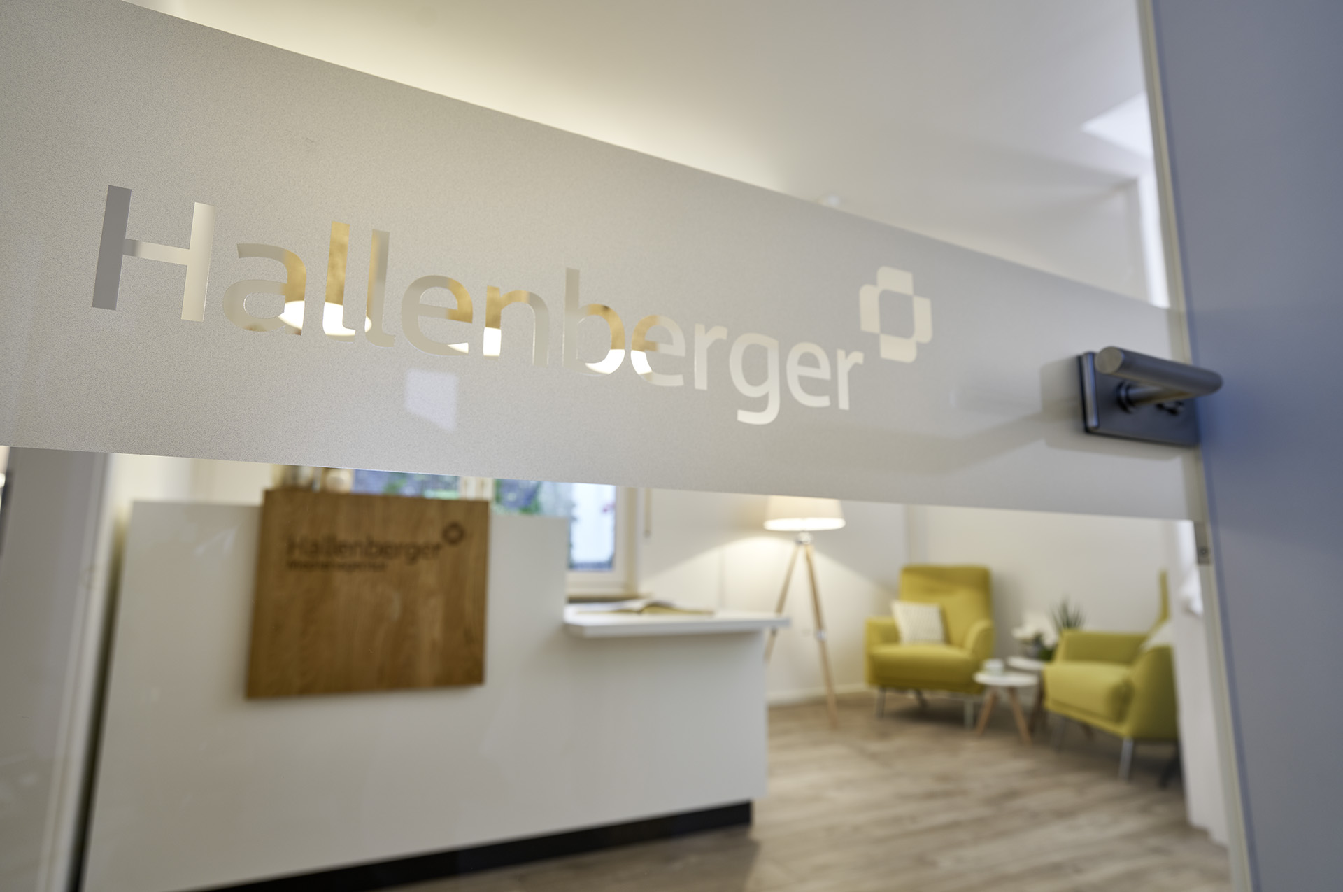 Eine Glastür mit einem Folienaufdruck "Hallenberger", durch die Tür sieht man verschwommen einen Schreibtisch, zwei grüne Sessel und eine Stehlampe.
