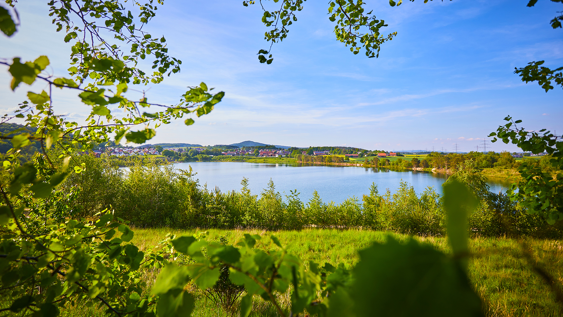 Ein See liegt umgeben von Natur, im Hintergrund sind Häuser sowie weitere Hügel, blauer Himmel mit wenig Wolken