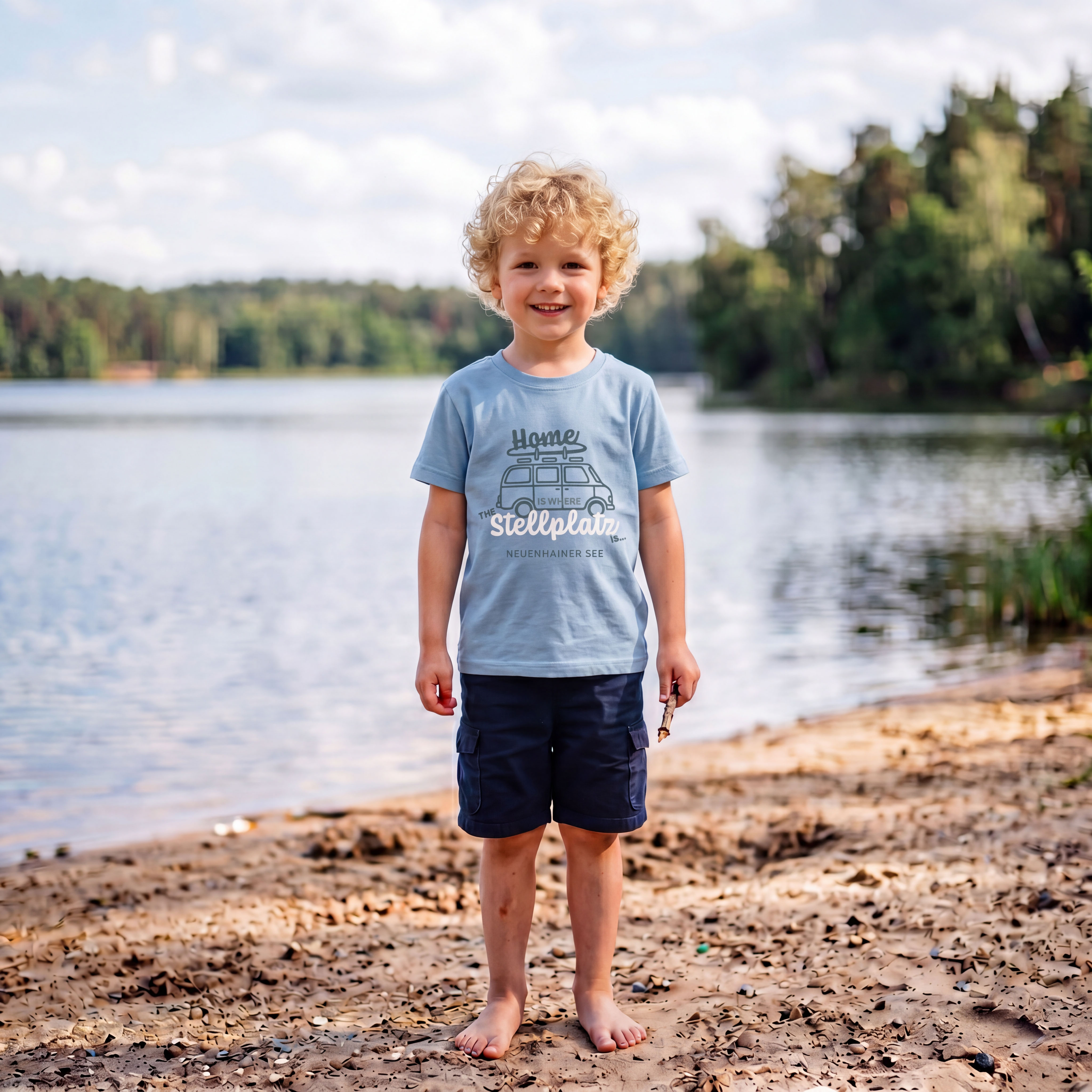 Ein Junge mit blonden Locken steht am Strand, im Hintergrund ein See und Bäume.