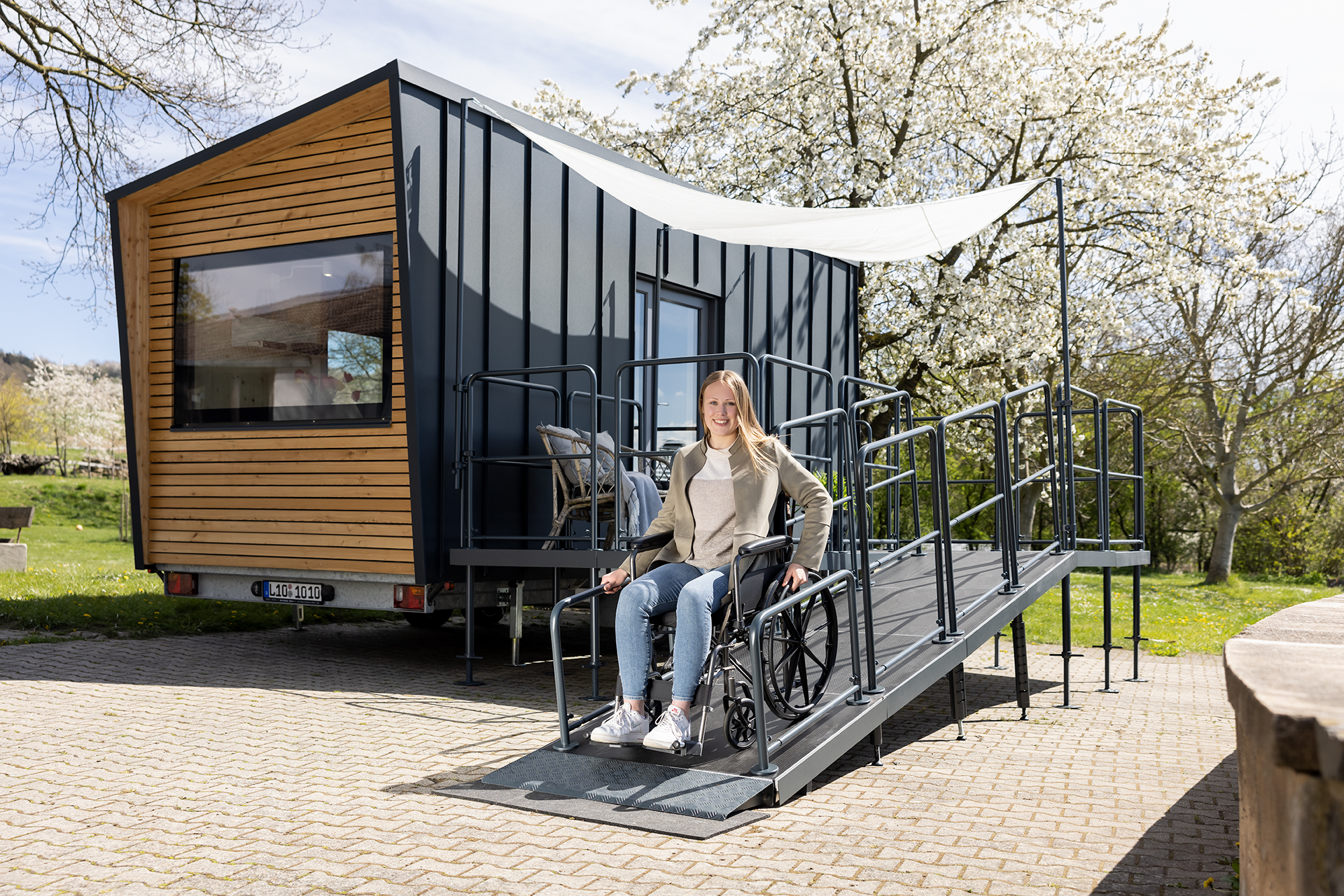 Ein barrierefreies Tiny House der Tischlerei Bock steht auf Asphalt und hat eine Rampe mit Terrasse angebaut, welche eine Rollstuhlfahrerin runterfährt